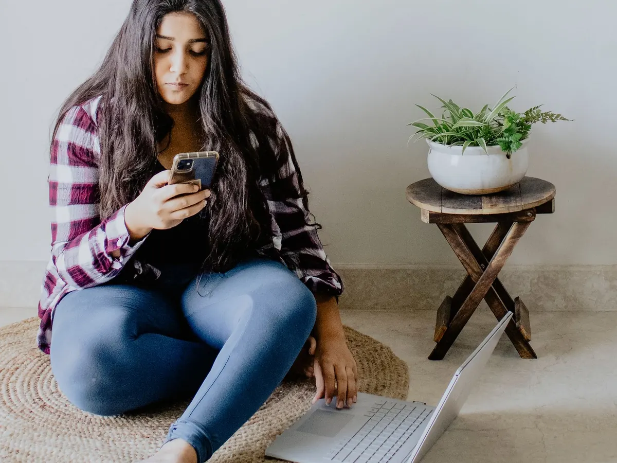 A young woman sitting on the floor of a bright room, looking at her smartphone with a laptop beside her